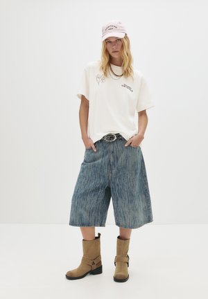 Une personne portant un t-shirt graphique blanc, un short en denim jusqu'aux genoux, des bottes beige à mi-mollet, une ceinture cloutée, et une casquette rose avec les mains dans les poches.