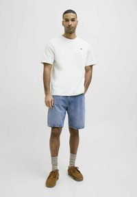 Camiseta blanca de manga corta con un pequeño acento rojo, combinada con shorts de mezclilla azules y zapatos de ante marrones. Calcetines grises con textura.