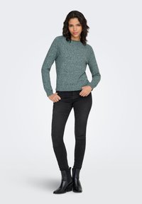 Šviesiai žalias tekstūruotas megztinis ilgomis rankovėmis, apvaliu kaklu ir ribuotu rankogaliu, derinamas su juodais siaurais džinsais ir juodais aulinukais.