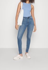 Top sin mangas de rayas en color azul claro, jeans ajustados desgastados en azul y zuecos gruesos blancos. La modelo lleva una bolsa tote blanca al hombro.