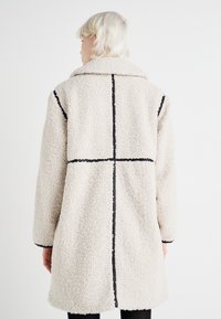 Beige, blødt, langt frakke med kravehals. Har sorte stikninger og en afslappet pasform.