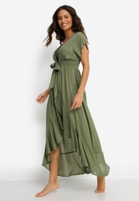 Μάξι φόρεμα σε ελαιί πράσινο, με λαιμόκοψη σε σχήμα V, κοντά μανίκια, ζώνη στη μέση και αέρινο ύφασμα με στρωμένο τελείωμα.