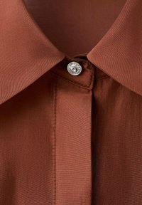 Cuello marrón de una camisa con una textura suave y satinada, adornado con un botón plateado decorativo y un diseño simple y limpio.