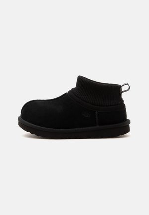 Zapato negro sin cordones con puño de punto en el tobillo, suela plana y tira en la parte trasera, diseñado para la comodidad y el uso cotidiano.