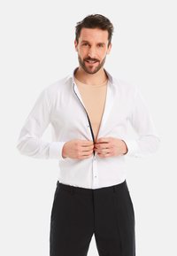 White long-sleeve shirt with a contrasting collar detail. Smooth fabric, button-up front, worn over a beige shirt, paired with black trousers.