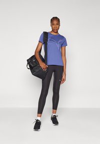 T-shirt roxo com logotipo branco, leggings pretas e mala preta. Modelo usa ténis pretos com detalhes brancos e verdes. Design e ajuste simples.