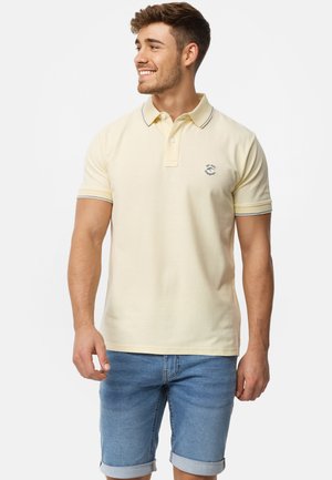 Polo-shirt i lysegul bomuld med krave, korte ærmer, blå trim og et lille logo på brystet. Båret med lyseblå denim shorts.