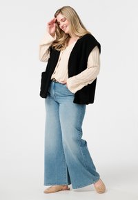 Zwarte getextureerde vest over een crèmekleurig geribd langemouwen shirt, gecombineerd met lichtblauwe broek met wijdere pijpen en tan kleurige geweven instap schoenen.