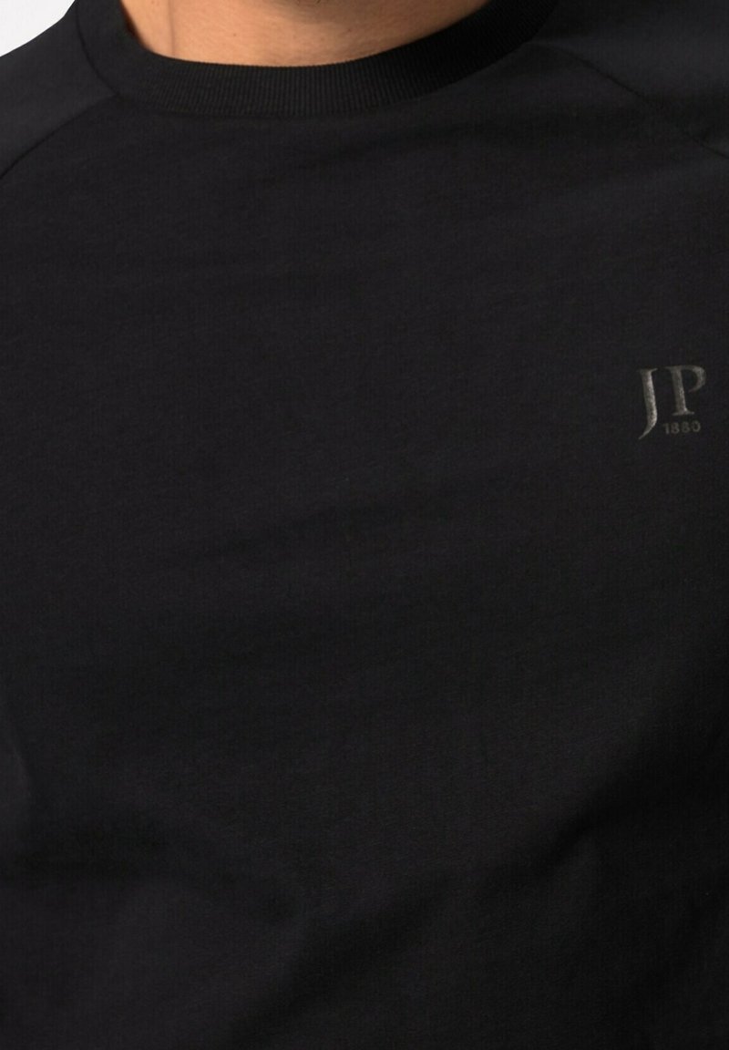 Homme portant un t-shirt noir à col rond avec un petit logo texte "JP 1880" sur le haut du côté droit de la poitrine.