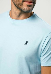 Vīrietis ar īsu bārdu, tērpies gaiši zilā T-kreklā ar mazu melnu silueta logotipu, kurā redzama staigājoša persona krūtīs.