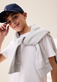 Grå sweatshirt lindad runt axlarna på en person, kombinerad med en vit t-shirt och en marinblå keps med en logotyp. Neutral bakgrund.