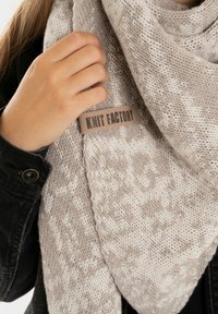 Lichtgrijze gebreide sjaal met een gestructureerd patroon en een bruine label met het opschrift "Knit Factory." De sjaal is casual gedragen over een donkere jas.