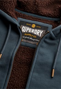 Marmoreret hættejakke med en mørk teal yderside, brun sherpa-foring og lynlåse i guldtoner. Har et sort mærke med teksten "Superdry Supply Co."