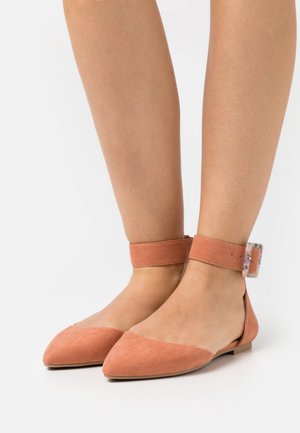 Béžové semišové baleríny s úzkou špičkou a širokými kotníkovými pásky s průhlednými obdélníkovými sponami na nahých nohách.
