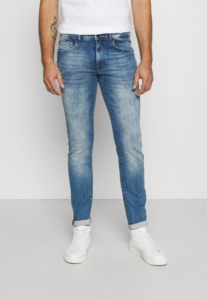 Valkoisen vaaleansinisen denim-farkut, joissa on haalistunut pesu, slim fit -leikkaus ja käännetyt lahkeet, yhdistettyinä valkoisiin korkeavartisiin tennareihin.
