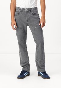 Pantalons en denim gris à coupe décontractée, avec poches à rabat et quincaillerie chromée, associés à des baskets bleues. Surface texturée.