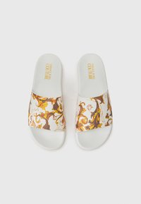 Hvite sandaler med en gul og brun barokk mønster over remmen. Gulltekst som leser "Versace Jeans Couture" på fotsengen.