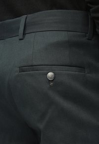 Calças cinza escuro apresentam uma textura lisa, um bolso traseiro com fecho de botão prateado e um cós bem arranjado.