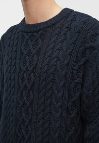 Pogled od blizu na osobu koja nosi tamnoplavu džemper s pletenim uzorakom i rebrastim ovratnikom.