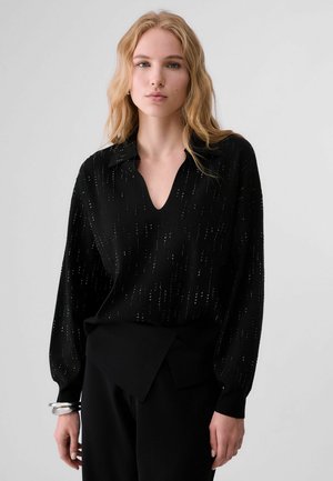 Giovane donna con lunghi capelli biondi, che indossa una blusa nera testurizzata a maniche lunghe e pantaloni neri su uno sfondo semplice.