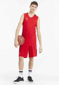 Rotes sportliches Tanktop und Shorts aus feuchtigkeitsableitendem Material, mit weißen Akzenten und dem Puma-Logo. Hält einen Basketball; schwarze Sneakers mit weißen Sohlen.