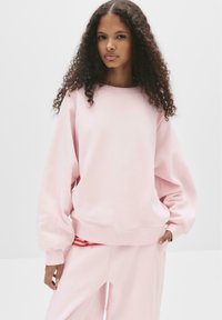 Felpa rosa chiaro con maniche lunghe, scollatura rotonda e vestibilità rilassata. Tessuto morbido con polsini e vita a coste. Strisce rosse visibili sotto.