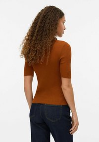 Femme avec de longs cheveux bouclés portant un haut à manches courtes en rib à rayures couleur rouille ajusté et un jean bleu foncé, se tenant de profil devant un fond uni.