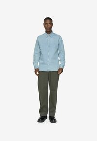 Lichtblauw denim overhemd met button-down kraag, lange mouwen en zichtbare stiksels, gecombineerd met olijfgroene broek en zwarte schoenen.