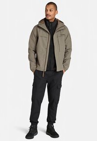 Chaqueta ligera de color verde oliva con capucha, cierre de cremallera y dos bolsillos delanteros; combinada con una capa interior negra con cremallera y pantalones cargo negros.