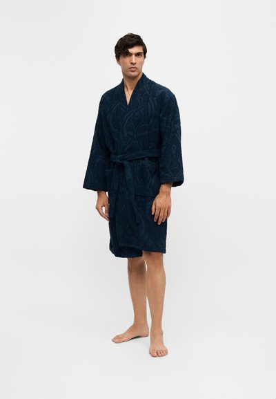Homme debout pieds nus portant un peignoir texturé bleu foncé noué à la taille sur un fond clair uni.