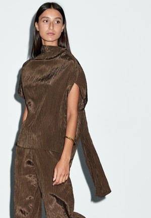 Top marrone plissettato con colletto alto e maniche corte, abbinato a pantaloni coordinati. Tessuto liscio e righe verticali. Accento di bracciale dorato.