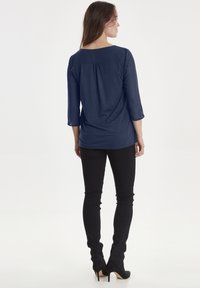Maglietta blu navy a maniche lunghe con scollatura quadrata, dettaglio arricciato sul retro, abbinata a jeans skinny neri e scarpe con tacco alto nere.
