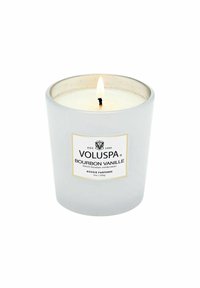 Vit cylindrisk ljus i en glasbehållare med en guldfärgad etikett. Har en tänd veke och läser "VOLUSPA BOURBON VANILLE." Kapacitet 9 oz.