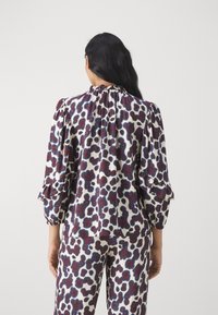 Blusa stampata con maniche arricciate; presenta una base bianca con motivi astratti in bordeaux e blu; il colletto arricciato aggiunge texture.