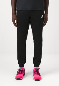Pantalones deportivos negros con un diseño ajustado, que cuentan con bolsillos laterales y un logo blanco bordado. Combinados con zapatillas deportivas de color rosa brillante.