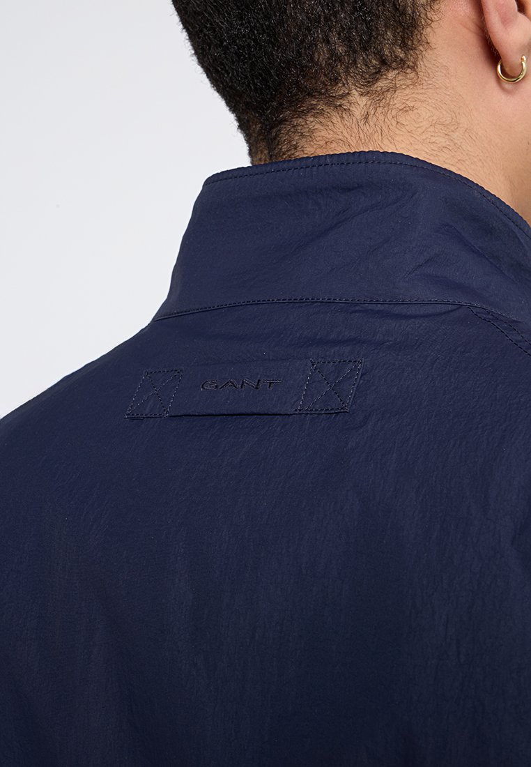 Marineblaue, leichte Jacke mit einer strukturierten Oberfläche. Verfügt über ein Rückenkragenetikett mit dem Markennamen "GANT" und auffälligen Stichakzenten.