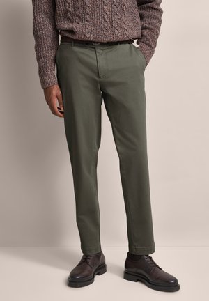 Olivgrüne Chinos mit entspannter Passform, mit flachem Bund, seitlichen Taschen und einem Gürtel, kombiniert mit dunkelbraunen Schuhen und einem braunen Strickpullover.