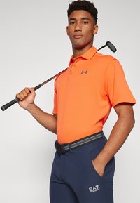 Polo shirt texturizado naranja con logo negro, combinado con pantalones marinos con un cinturón a rayas y un pequeño logo blanco. Sosteniendo un palo de golf.