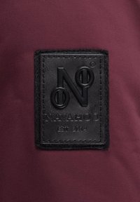 Dieprode stof heeft een rechthoekige zwarte leren patch met een reliëflogo "N" en "NAVAHOO EST 2014," dat textuurdetails toont.