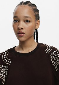 Hanorac maro cu detalii embellisate pe umeri, decorate cu mărgele și broderie. Material moale, decolteu rotund și croială lejeră.