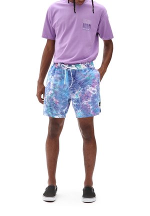 Man die een paarse Vans T-shirt draagt, blauwe en paarse tie-dye short met witte trekkoord, en zwarte instapschoenen, staand met handen in de zakken.