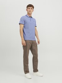 Blauwe polo met korte mouwen en een zwarte kraag, gecombineerd met bruine broek en witte sneakers. De stof lijkt gestructureerd en casual.