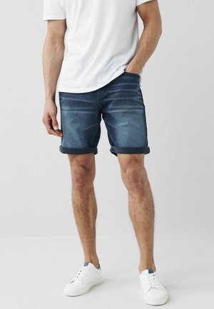 Szorty dżinsowe w ciemnym niebieskim kolorze z efektami sprania; podwinięte mankiety, pięciokieszeniowy fason, zestawione z białymi trampkami i gładką białą koszulką.