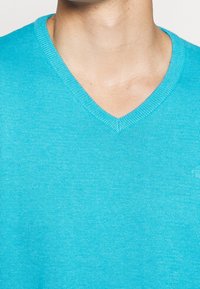 Pull à col en V turquoise vif, fabriqué en tissu tricoté léger. Présente une texture subtile avec un petit détail de logo sur le côté.