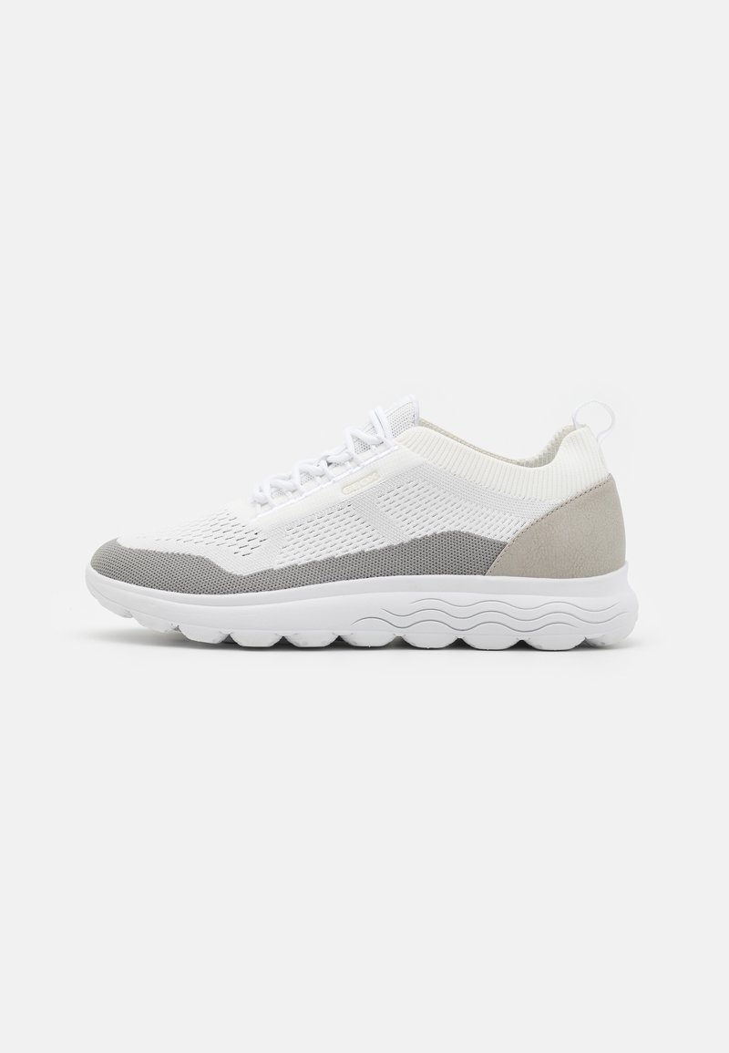 Zapatilla deportiva de corte bajo en blanco y gris con parte superior de punto, frente con cordones, cuello acolchado y suela blanca texturizada con patrón en forma de ola.