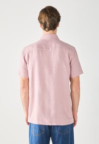 Camisa de manga corta en tela rosa clara con textura suave. Presenta un cuello clásico, costura en el yugo trasero y un corte relajado.