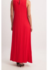 Robe longue rouge en tissu texturé, sans manches, avec un détail dos en forme de goutte et une silhouette fluide. Portée avec des talons noirs.