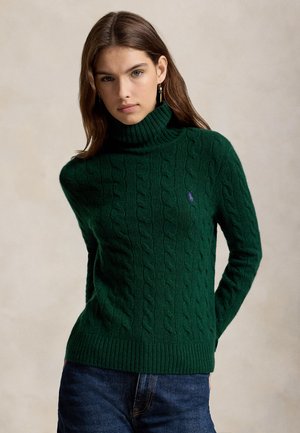 Jeune femme aux longs cheveux bruns portant un pull à col roulé torsadé vert foncé et un jean bleu sur un fond neutre.