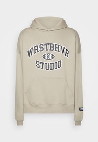 Beige capuchontrui met voorzak. Heeft een grote zwarte grafische opdruk "WRSTBHVRA STUDIO" en geribbelde manchetten en zoom.