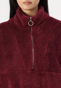 Burgunderfarget fleece-pullover med høy krage, frontlukking med glidelås og teksturert stoffmønster. Merkbar sølvfarget glidelåskomponent.